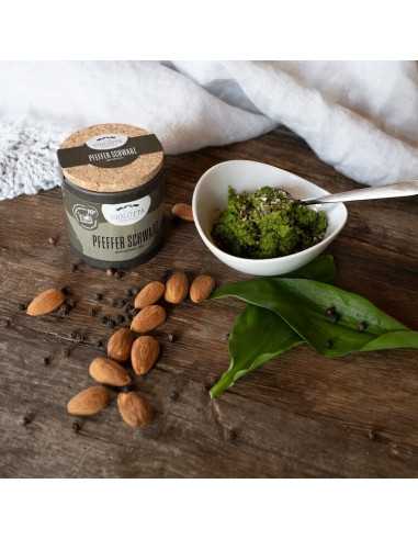 Szene auf Holzuntergrund mit Pfefferdose, Mandeln, Bärlauchblättern und einer Schale grünes Pesto.