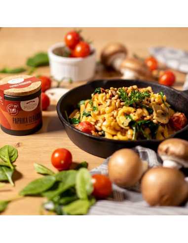 Schwarze Pfanne mit buntem Nudelgericht auf Holzplatte, daneben die Paprika-Gewürzdose, Pilze, Tomaten und Spinatblätter.