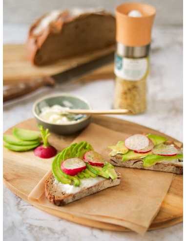 Zwei belegte Brotscheiben mit Avocado, Radieschen und Dip auf Holzbrett, dahinter die Gewürzmühle mit grünem Etikett.