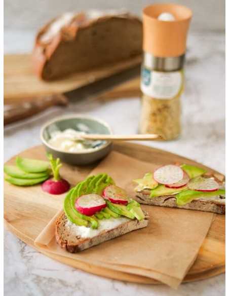 Zwei belegte Brotscheiben mit Avocado, Radieschen und Dip auf Holzbrett, dahinter die Gewürzmühle mit grünem Etikett.