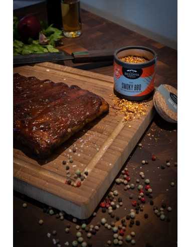 Geöffnete Pappdose mit Smoky BBQ Rub neben mariniertem Grillfleisch auf einem Holzbrett mit buntem Pfeffer