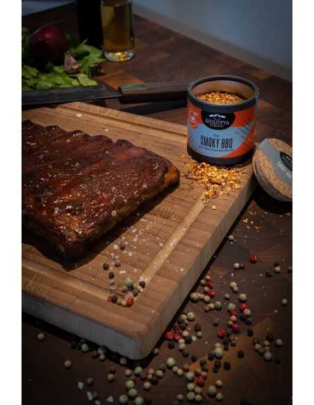 Geöffnete Pappdose mit Smoky BBQ Rub neben mariniertem Grillfleisch auf einem Holzbrett mit buntem Pfeffer