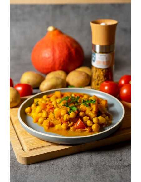 Teller mit Currygericht aus Kichererbsen und Gemüse; Kürbis, Kartoffeln, Tomaten und die Gewürzmühle mit orangem Etikett.