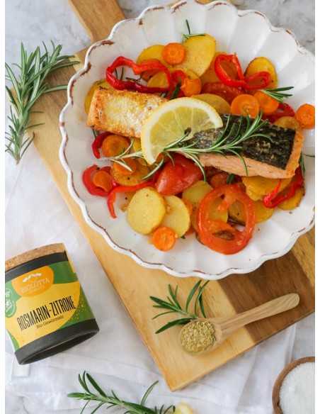 Schwarze Pappdose neben einem Teller mit Lachsfilet, Zitronenscheibe, Rosmarin und buntem Ofengemüse