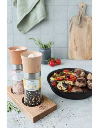 Zwei Gewürzmühlen mit Holzaufsatz in Halterung aus Holz, im Hintergrund bunter Gemüseteller mit Fleischbällchen