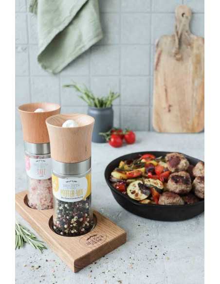 Zwei Gewürzmühlen mit Holzaufsatz in Halterung aus Holz, im Hintergrund bunter Gemüseteller mit Fleischbällchen