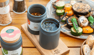food_mobil-1000 Verschiedene Biolotta-Gewürzprodukte auf einem Holztisch, darunter geöffnete Keramiktöpfchen mit Chili und Öl sowie gegrilltes Gemüse auf einem Teller im Hintergrund.