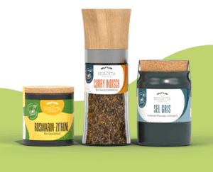 BioLotta Gewuerze_Darstellung Drei Biolotta-Gewürzprodukte auf grünem Untergrund: ein gelbes Töpfchen „Rosmarin-Thymian“, eine Gewürzmühle „Curry Indisch“ und ein Keramiktöpfchen „Sel Gris“ mit Korkdeckel.