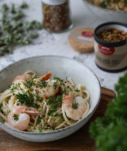 Ein Teller mit cremiger Pasta und Garnelen, garniert mit frischer Petersilie. Im Hintergrund sind eine Dose Knoblauch-Chili-Gewürz und frische Kräuter zu sehen. Das Gericht ist appetitlich angerichtet und versprüht italienischen Charme.