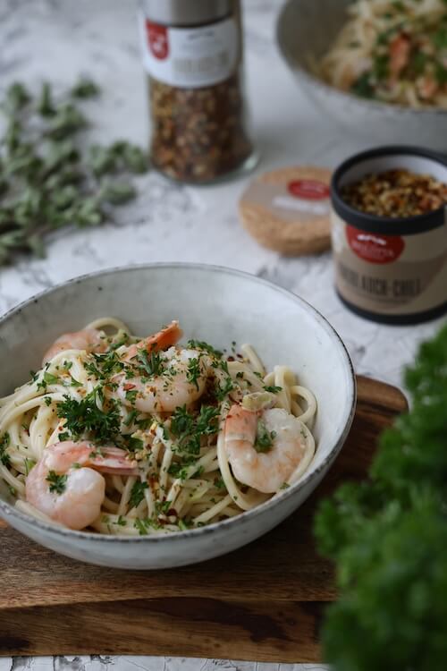 Ein Teller mit cremiger Pasta und Garnelen, garniert mit frischer Petersilie. Im Hintergrund sind eine Dose Knoblauch-Chili-Gewürz und frische Kräuter zu sehen. Das Gericht ist appetitlich angerichtet und versprüht italienischen Charme.