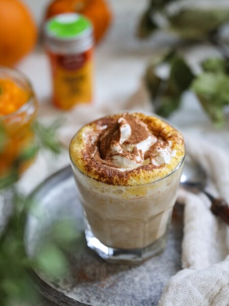 Glas mit cremigem Gewürz-Kürbis-Latte, getoppt mit Sahne und Pumpkin Spice, auf einem silbernen Tablett mit herbstlicher Dekoration im Hintergrund.
