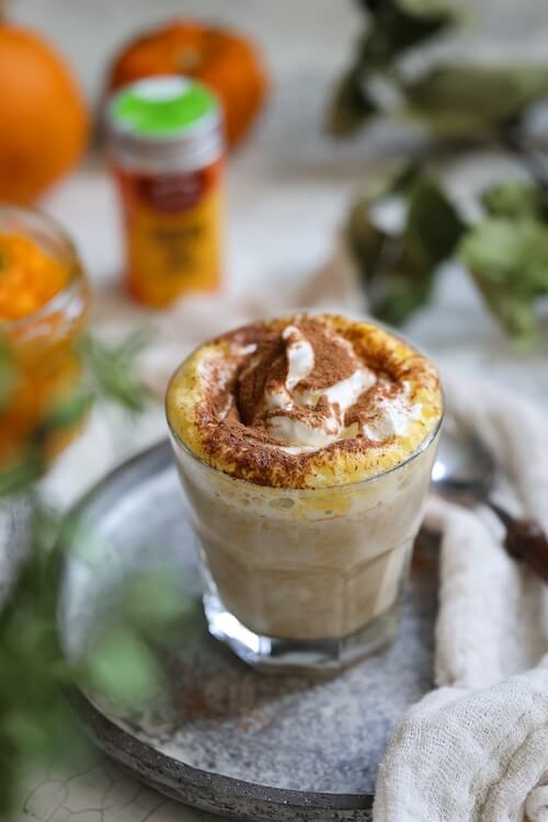 Glas mit cremigem Gewürz-Kürbis-Latte, getoppt mit Sahne und Pumpkin Spice, auf einem silbernen Tablett mit herbstlicher Dekoration im Hintergrund.
