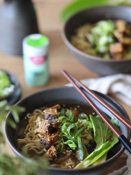 Zwei Schalen mit dampfender Umami-Suppe, belegt mit knusprigem Tofu, Pak Choi, Frühlingszwiebeln und Sesam – serviert mit Essstäbchen auf rustikalem Holztisch.