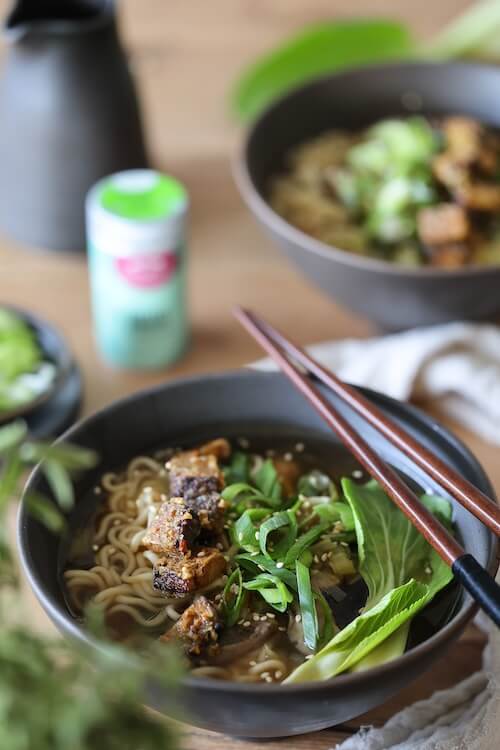 Zwei Schalen mit dampfender Umami-Suppe, belegt mit knusprigem Tofu, Pak Choi, Frühlingszwiebeln und Sesam – serviert mit Essstäbchen auf rustikalem Holztisch.