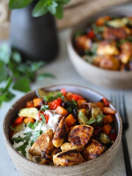 Nahaufnahme einer Hähnchenbowl mit goldbraun gebratenem Hähnchen, buntem Ofengemüse, Bulgur und Joghurt-Zitronen-Dip in einer rustikalen Schüssel, garniert mit frischer Minze.
