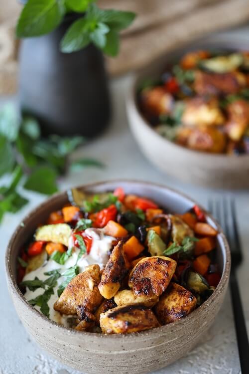 Nahaufnahme einer Hähnchenbowl mit goldbraun gebratenem Hähnchen, buntem Ofengemüse, Bulgur und Joghurt-Zitronen-Dip in einer rustikalen Schüssel, garniert mit frischer Minze.
