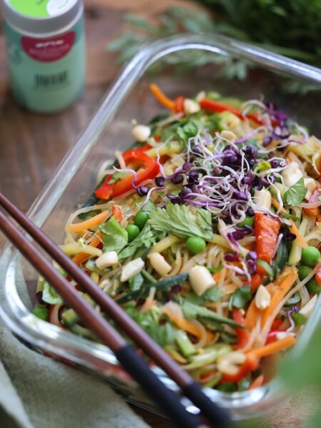 Asia-Glasnudelsalat mit buntem Gemüse, frischen Kräutern und Erdnüssen in Glasform, daneben ein Umami-Gewürzstreuer.