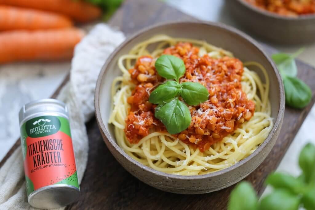 Vegane Linsen-Bolognese mit Spaghetti, garniert mit Basilikum, im Keramikschälchen, daneben italienische Kräutermischung von BioLotta.